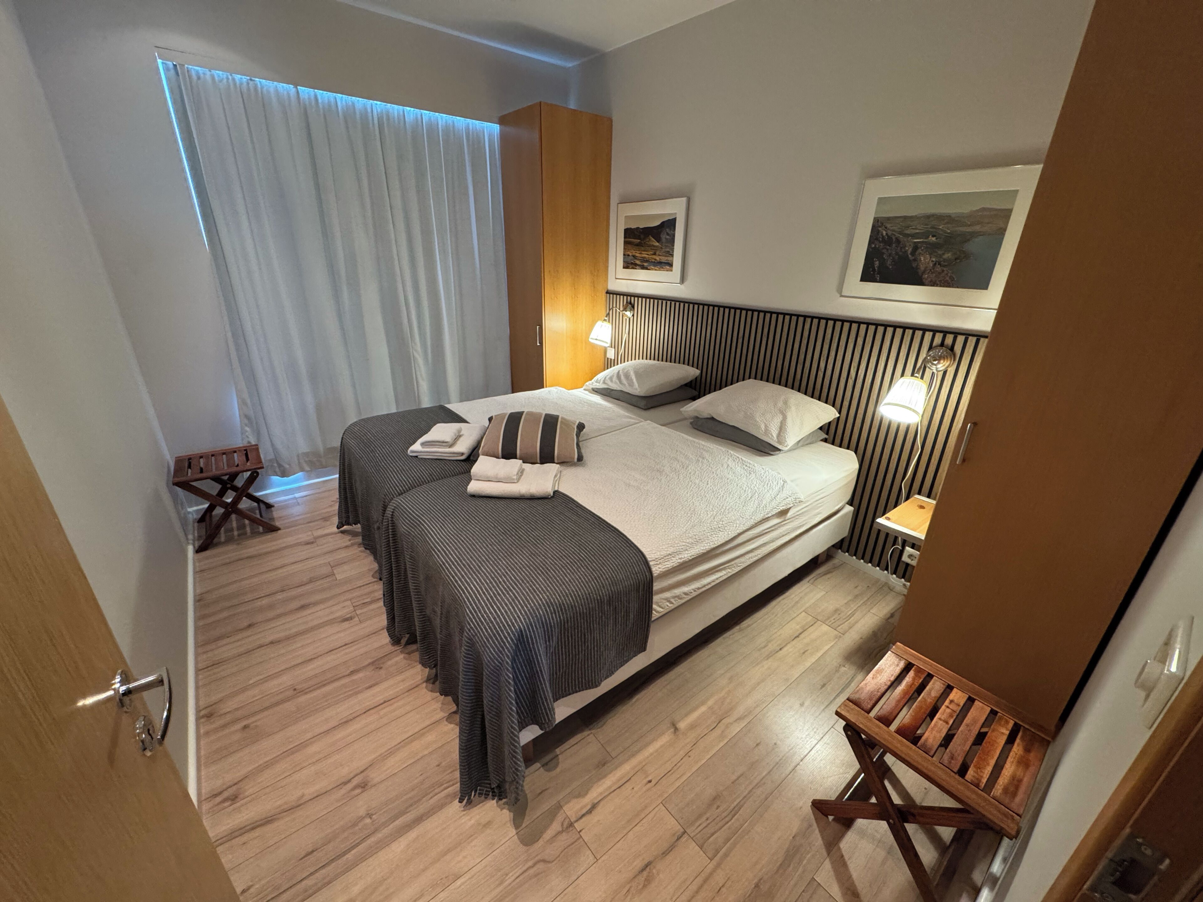 Una moderna camera d'albergo con un letto grande, due comodini, uno sgabello di legno e una finestra con tende.