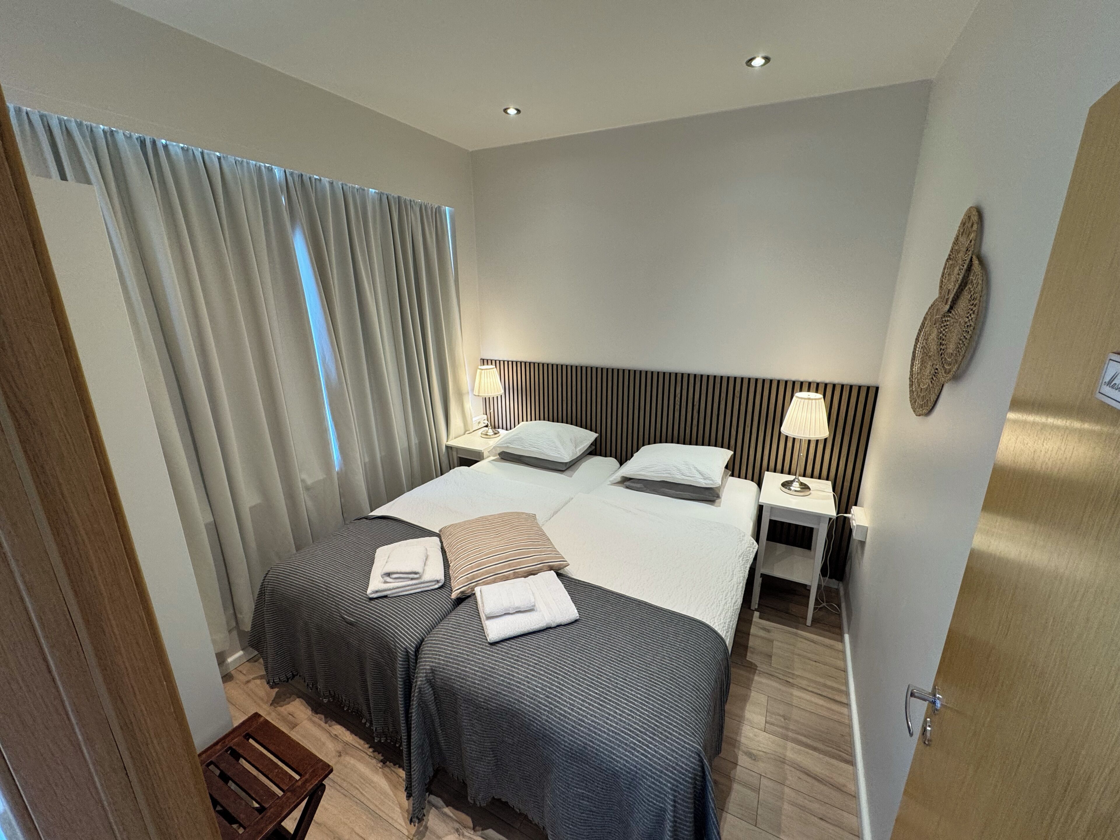 Et hotelværelse med to senge, en træbænk, et lille bord med en lampe og et gardin.