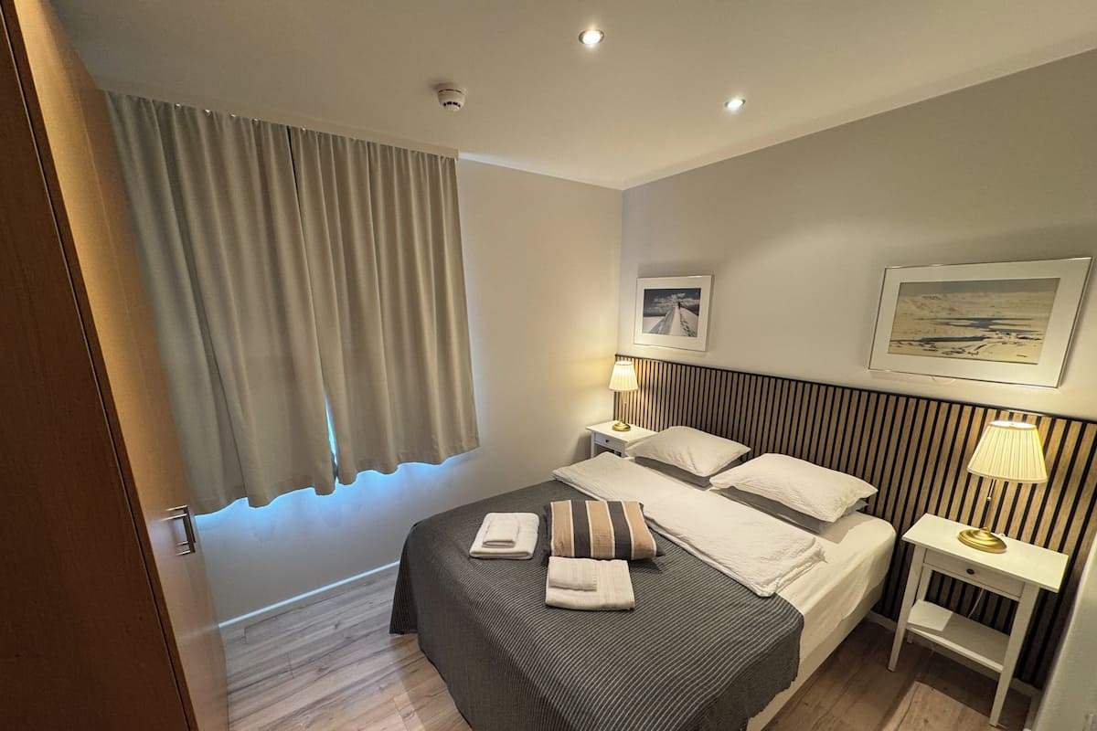 A hotel room with a bed, two bedside tables, a headboard, a window with curtains, and a wall-mounted picture.