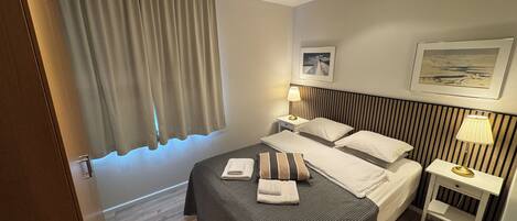 A hotel room with a bed, two bedside tables, a headboard, a window with curtains, and a wall-mounted picture.