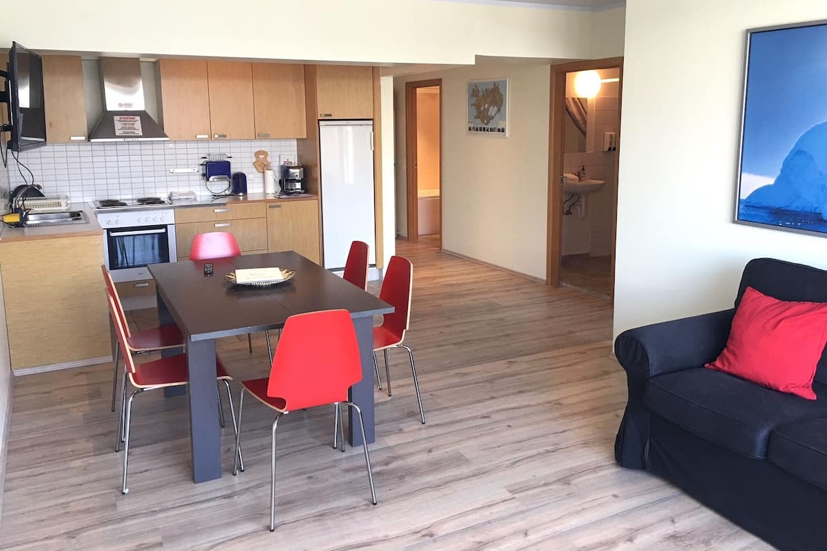 A modern kitchen with wooden cabinets, a dining table with red chairs, and a black sofa with a red pillow.