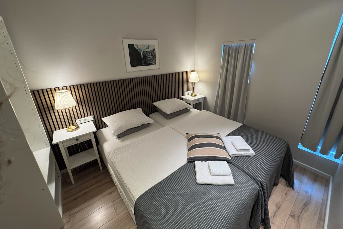 A hotel room with a bed, two bedside tables, a nightstand, a lamp, a window with curtains, and a framed picture on the wall.