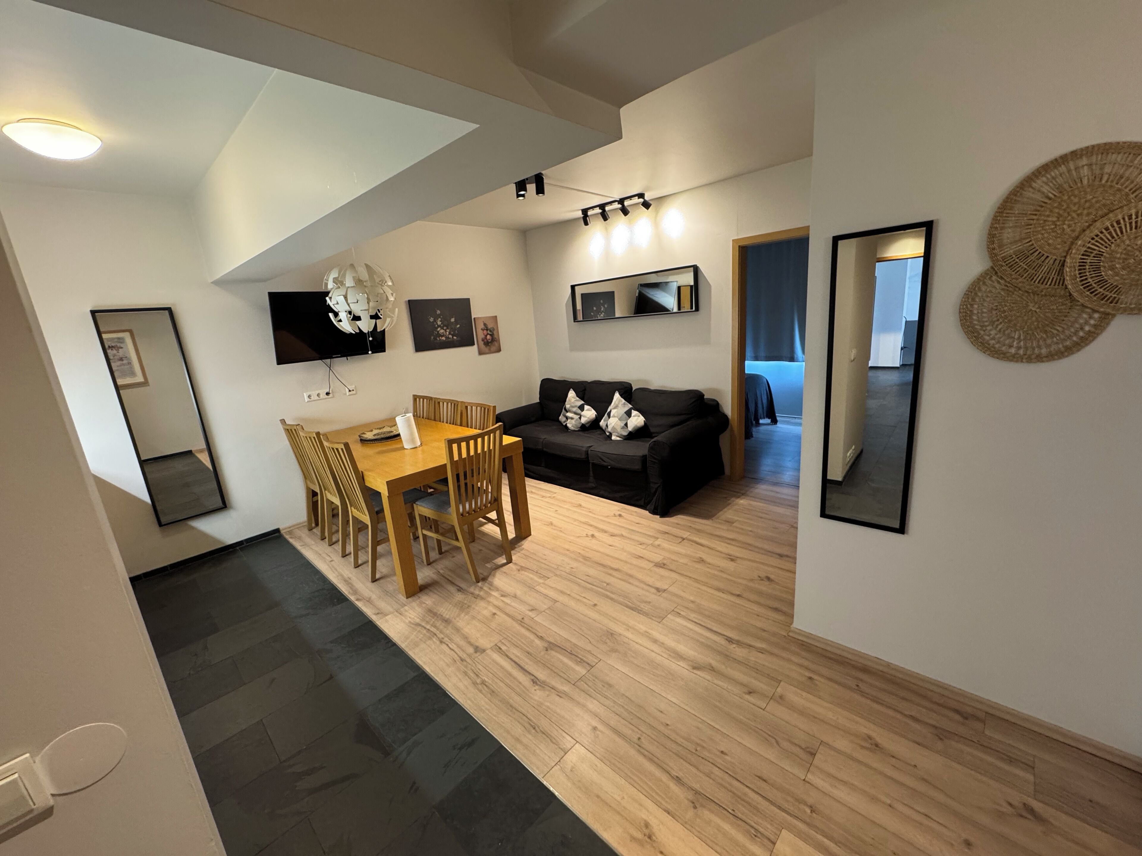 A modern living room with a dining area, a black sofa, and a TV mounted on the wall.