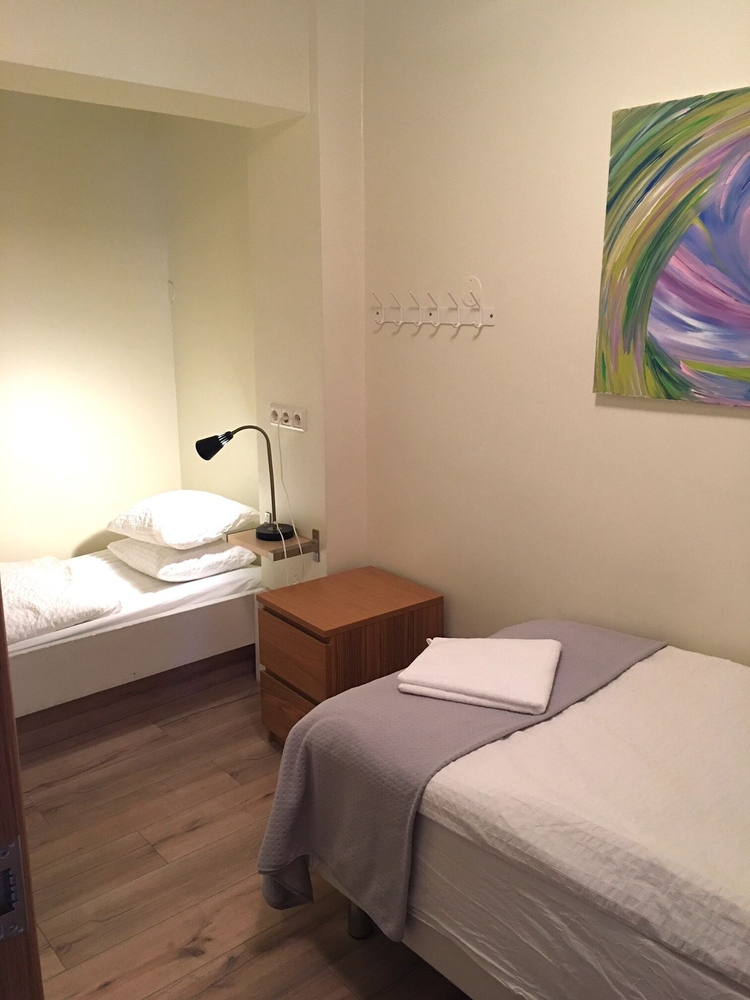 A bedroom with a bed, a bedside table, a lamp, a wall-mounted coat rack, and a painting.