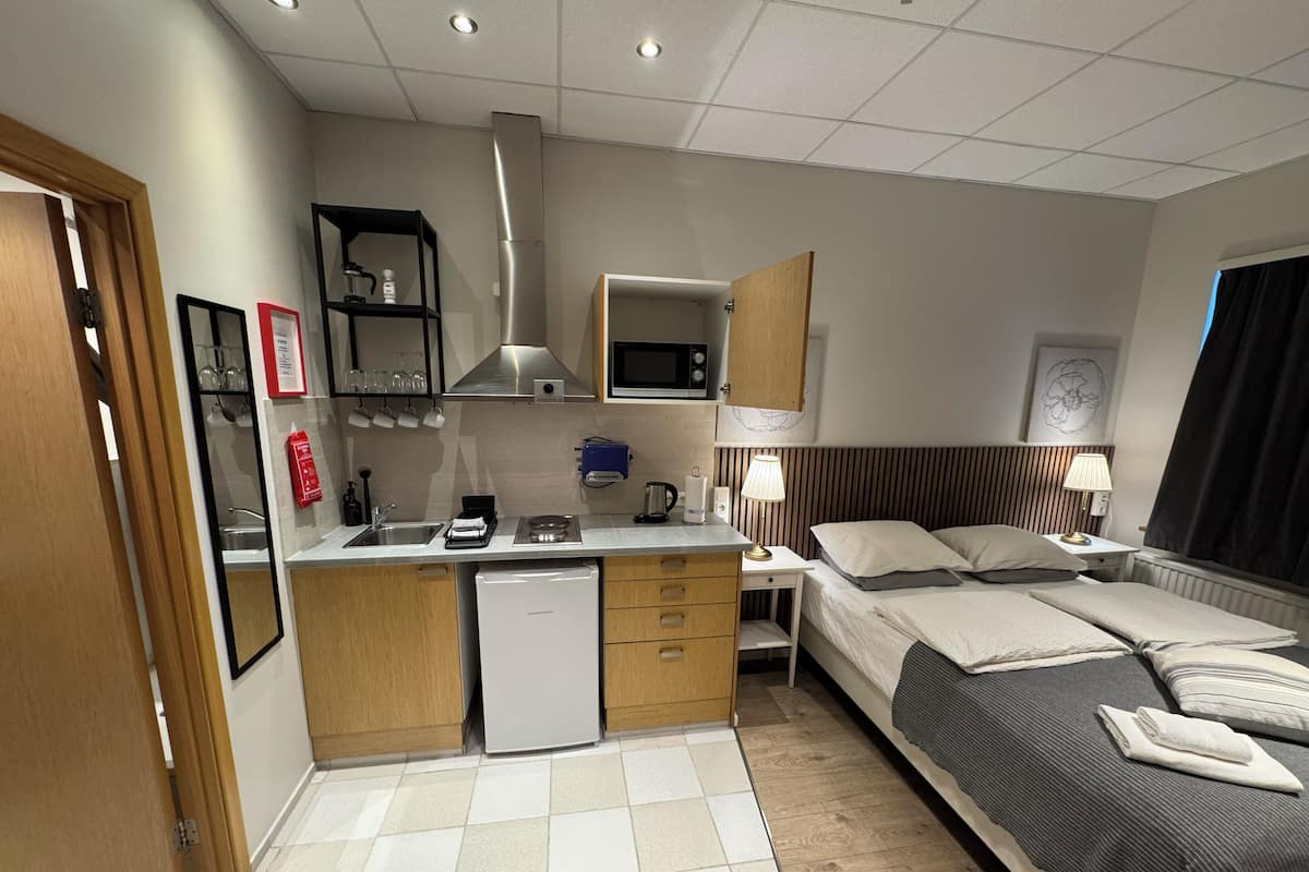 A compact hotel room with a kitchenette, a bed, bedside tables, and a window with curtains.