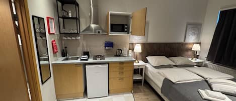 Una habitación de hotel compacta con kitchenette, una cama, mesitas de noche y una ventana con cortinas.