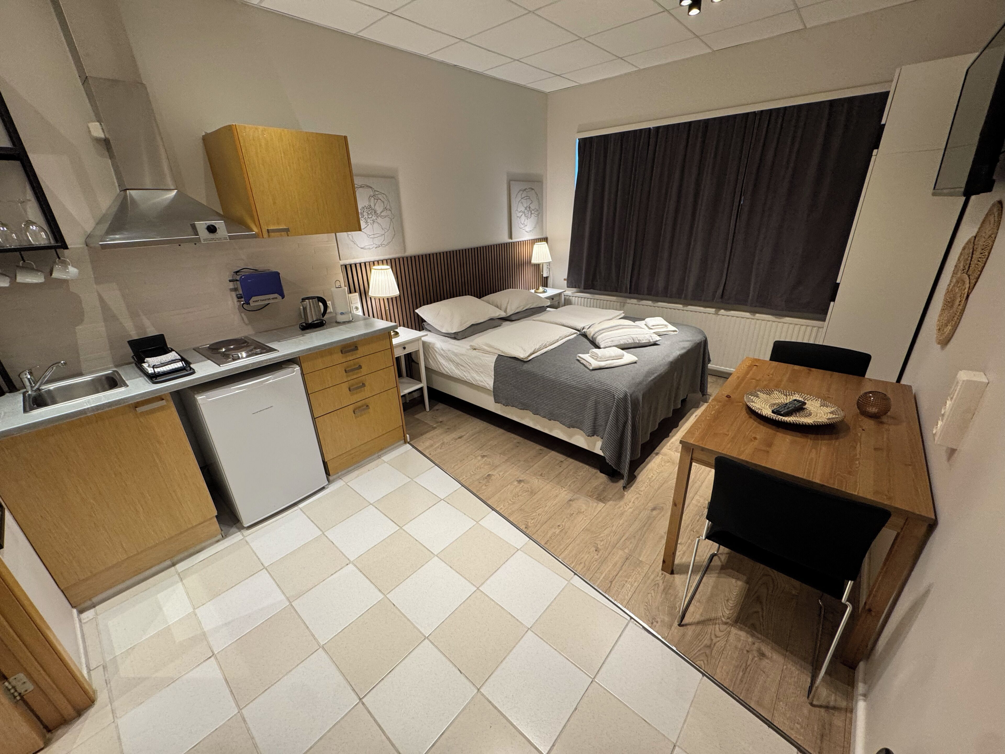 Ein kompaktes Hotelzimmer mit Küchenzeile, einem Bett mit Kopfteil, einem Esstisch und einem großen Fenster.
