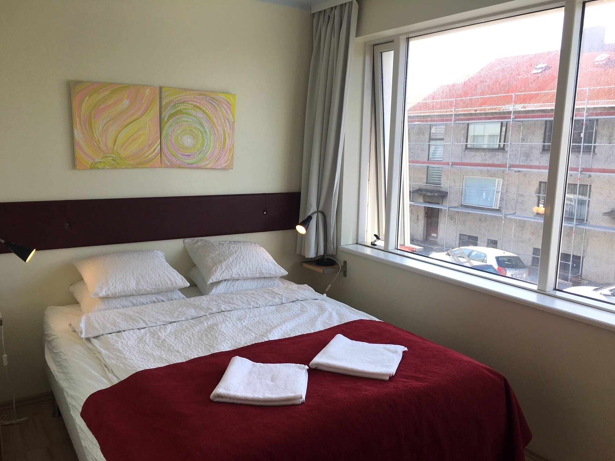 A hotel room with a bed, two pillows, a red blanket, and two abstract paintings on the wall.