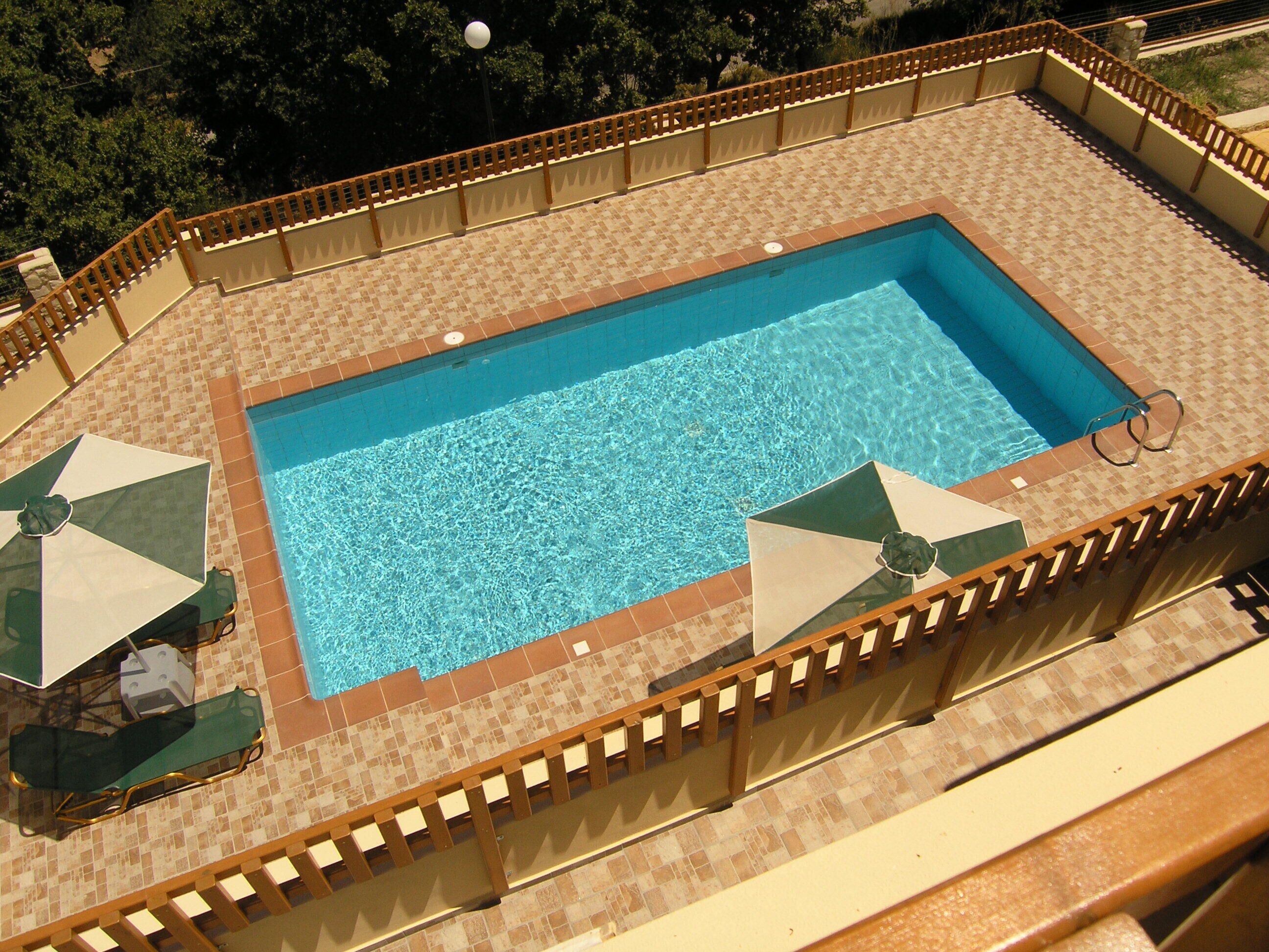A rectangular outdoor swimming pool with a tiled deck, surrounded by a wooden fence. There are two umbrellas and lounge chairs on the deck.