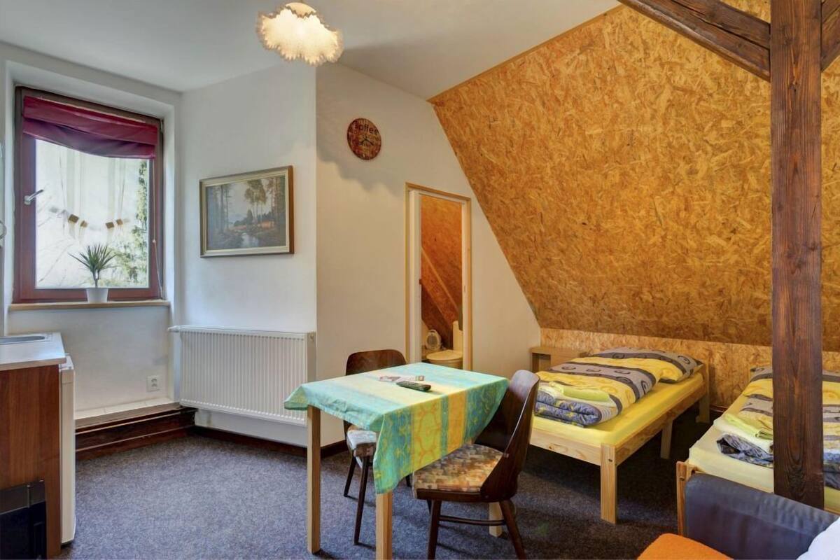 Una habitación pequeña y acogedora con techo inclinado, dos camas individuales, una mesa de madera con mantel colorido, una silla, un radiador, una ventana con cortina roja y un reloj en la pared.