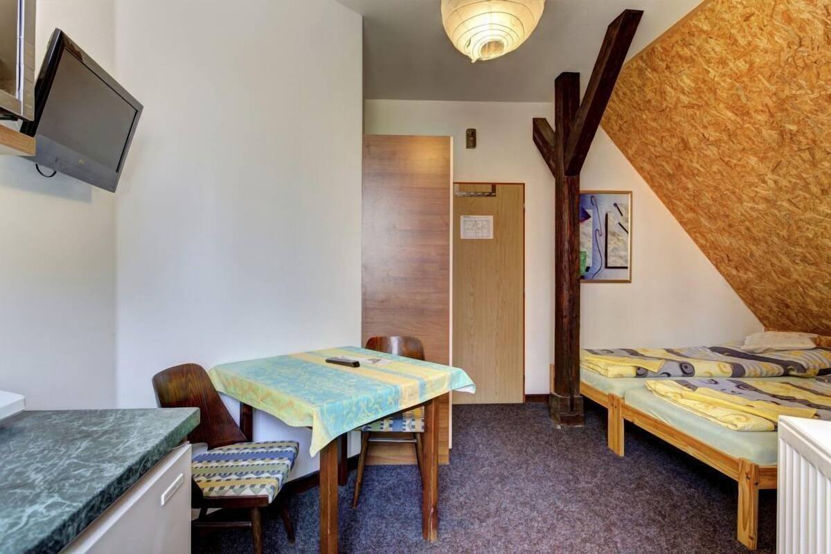 Una habitación pequeña con una cama, una mesita con una silla, un televisor en la pared y techo de madera.