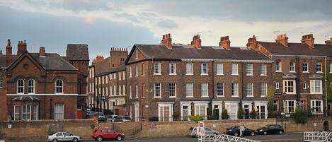 A row of red brick townhouses with white window frames, a canal in the foreground, and a pedestrian bridge.