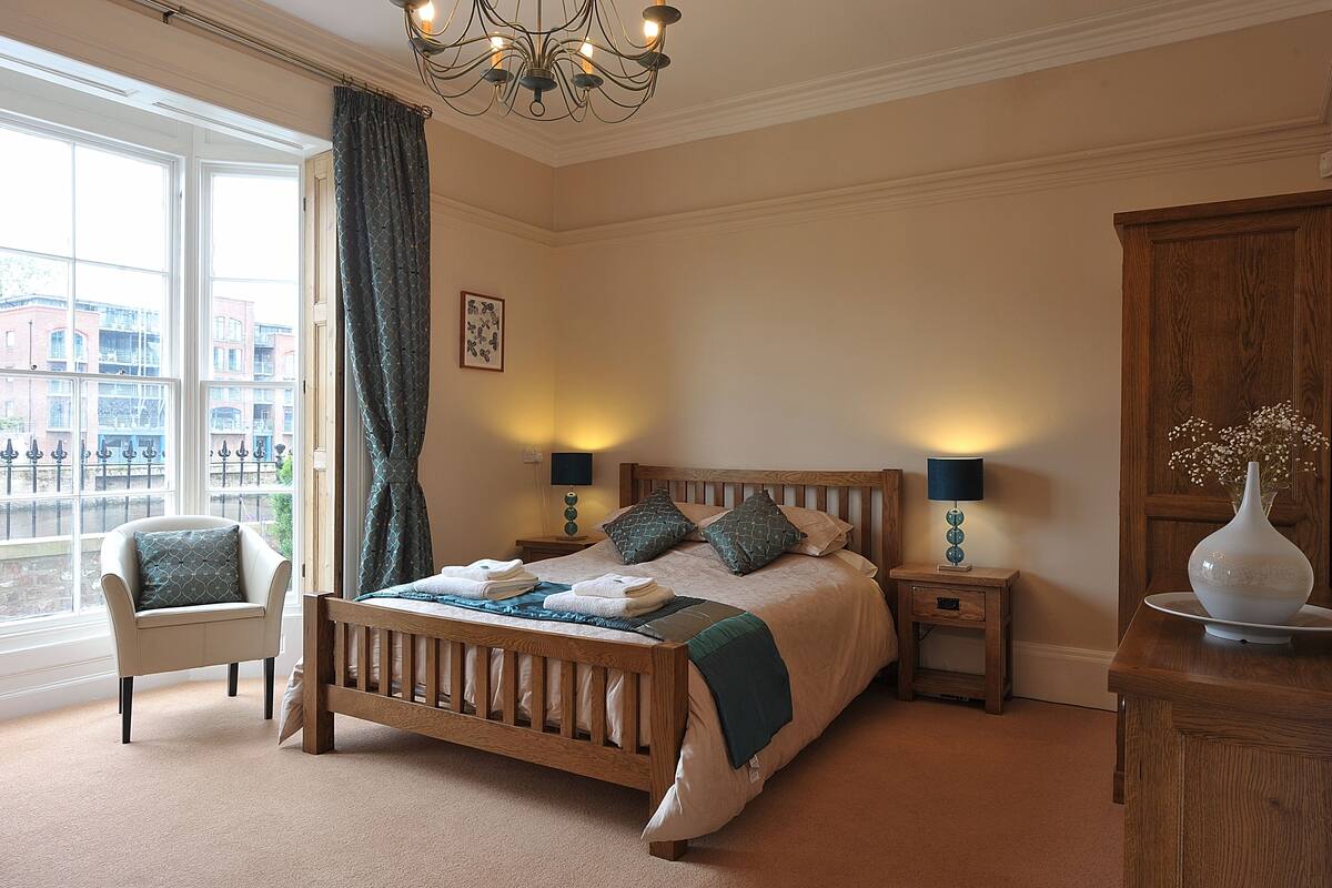 A bedroom with a wooden bed, a chair, a nightstand with lamps, and a view of buildings through large windows.