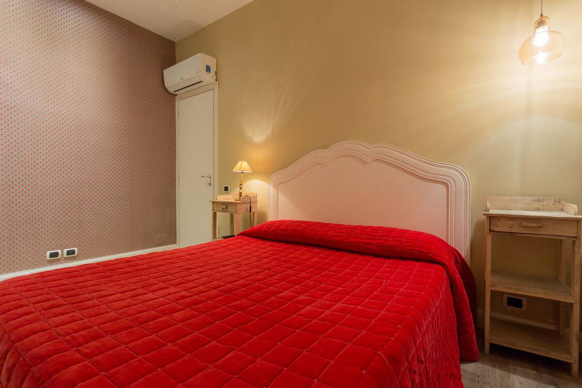 Un dormitorio con una colcha acolchada roja, cabecera blanca, mesita de noche con lámpara y aire acondicionado de pared.
