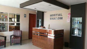 A reception desk with a sign reading "HOSTAL SHALOM" in a hotel lobby with a tiled floor, striped chairs, and a glass display cabinet. - Hostal Shalom (Pisco)