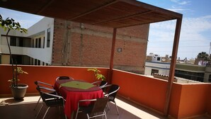 A covered outdoor seating area with a table and chairs, potted plants, and a view of buildings in the background. - Hostal Shalom (Pisco)