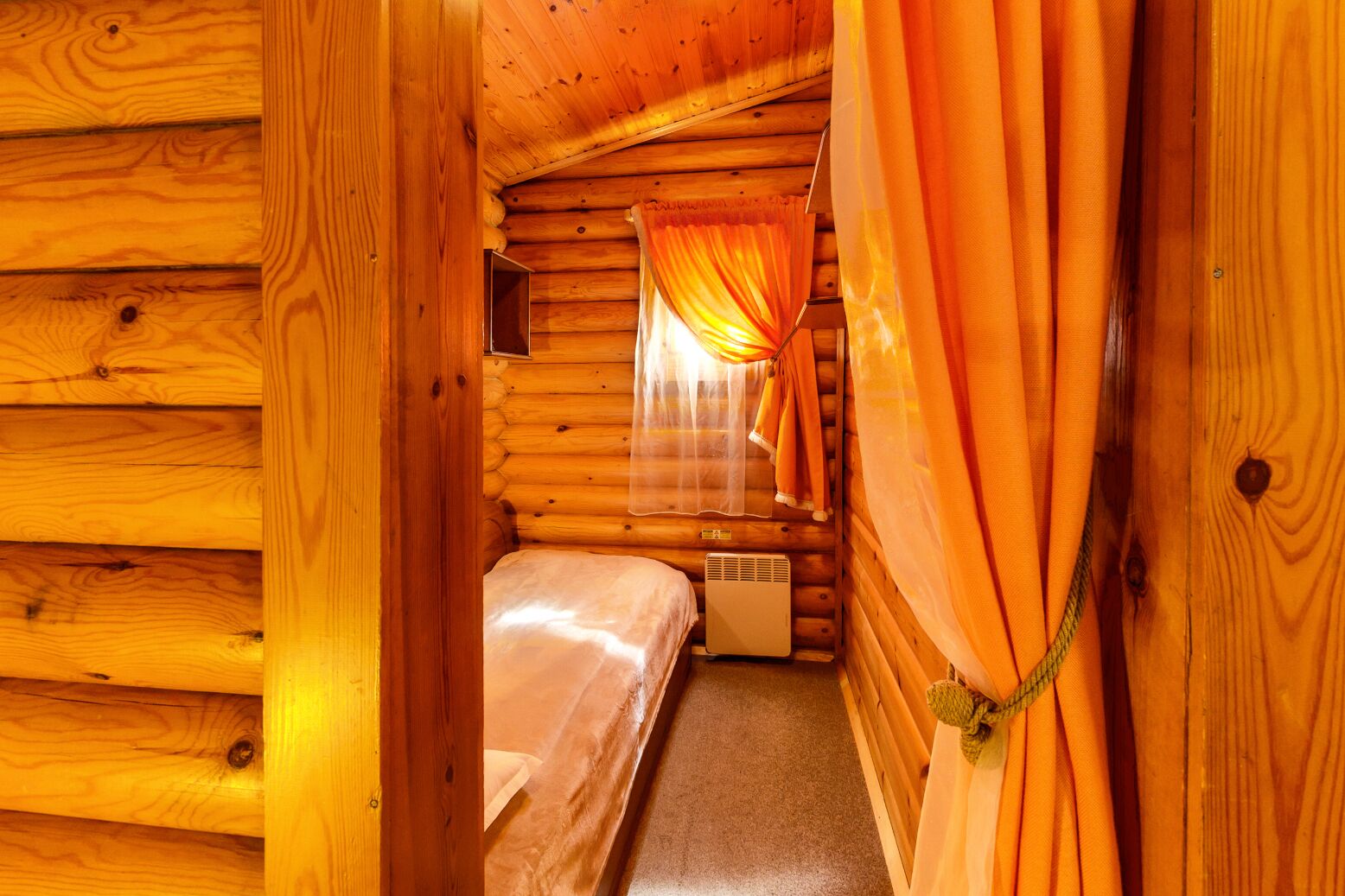 A wooden cabin room with a bed, a heater, and orange curtains.