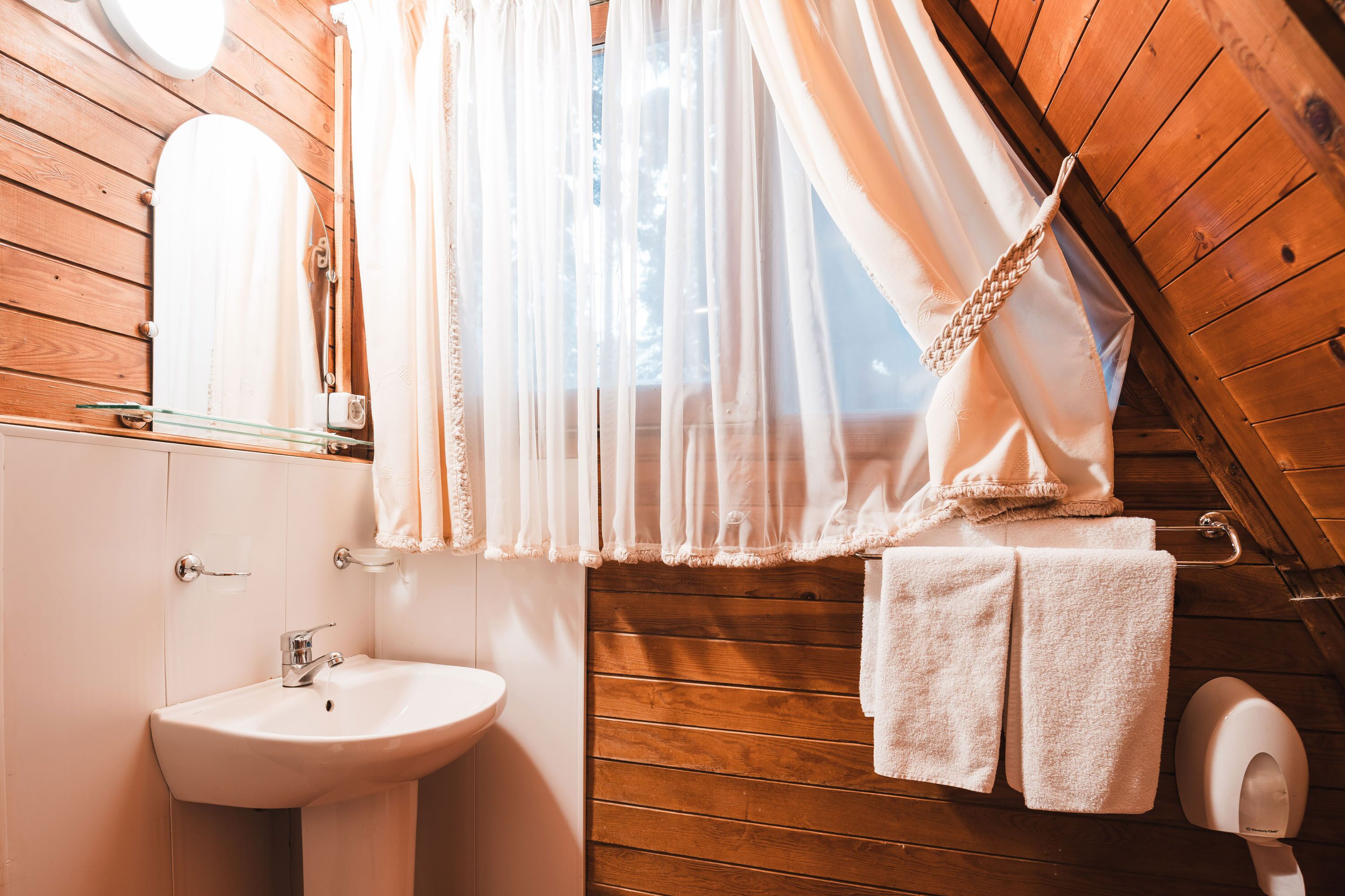 A wooden cabin bathroom with a white sink, a mirror, and a window with sheer curtains.