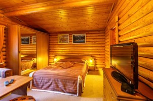 Quarto de cabana de tronco com uma cama, uma TV, uma escrivaninha com um computador e um espelho.