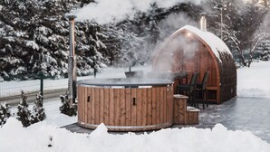 Un bain à remous en bois, doté d’une cheminée d’où s’échappe de la vapeur, entouré d’arbres couverts de neige et d’un chalet en bois.