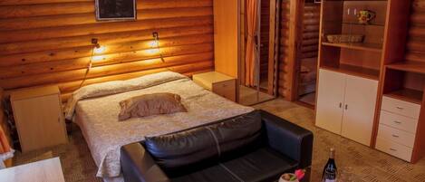 Acogedor dormitorio de cabaña de madera con cama, sofá de cuero, armario y una mesita con una botella y vasos.