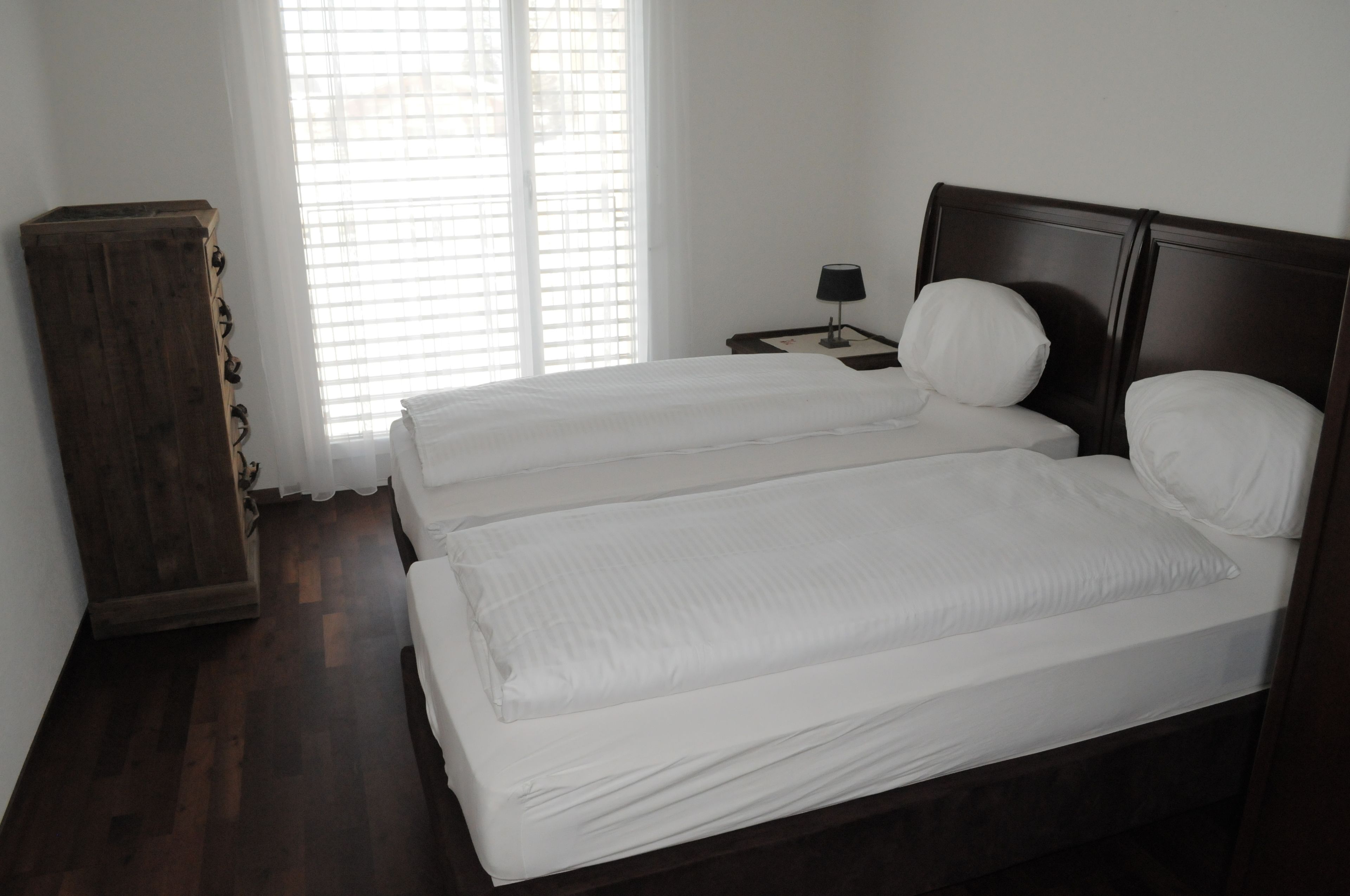 A double bed with white bedding, a wooden headboard, a bedside table with a lamp, and a window with blinds.