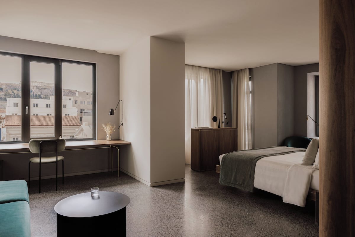A modern hotel room with a large window, a desk, a chair, a bed, and a round coffee table.