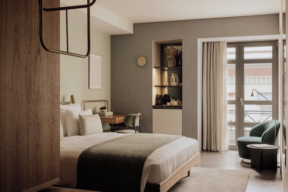 A modern hotel room with a large bed, a desk, a chair, a small table, and a balcony with curtains.