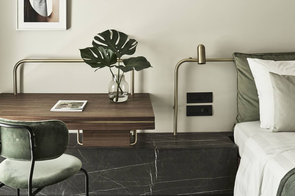A modern hotel room with a wooden desk, a green chair, a large plant, and a bed with a green pillow.