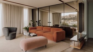 A modern living room with a large sofa, a small armchair, and a coffee table. There is a large window with a view of a cityscape. - Perianth Hotel, a member of Design Hotels (Athens)