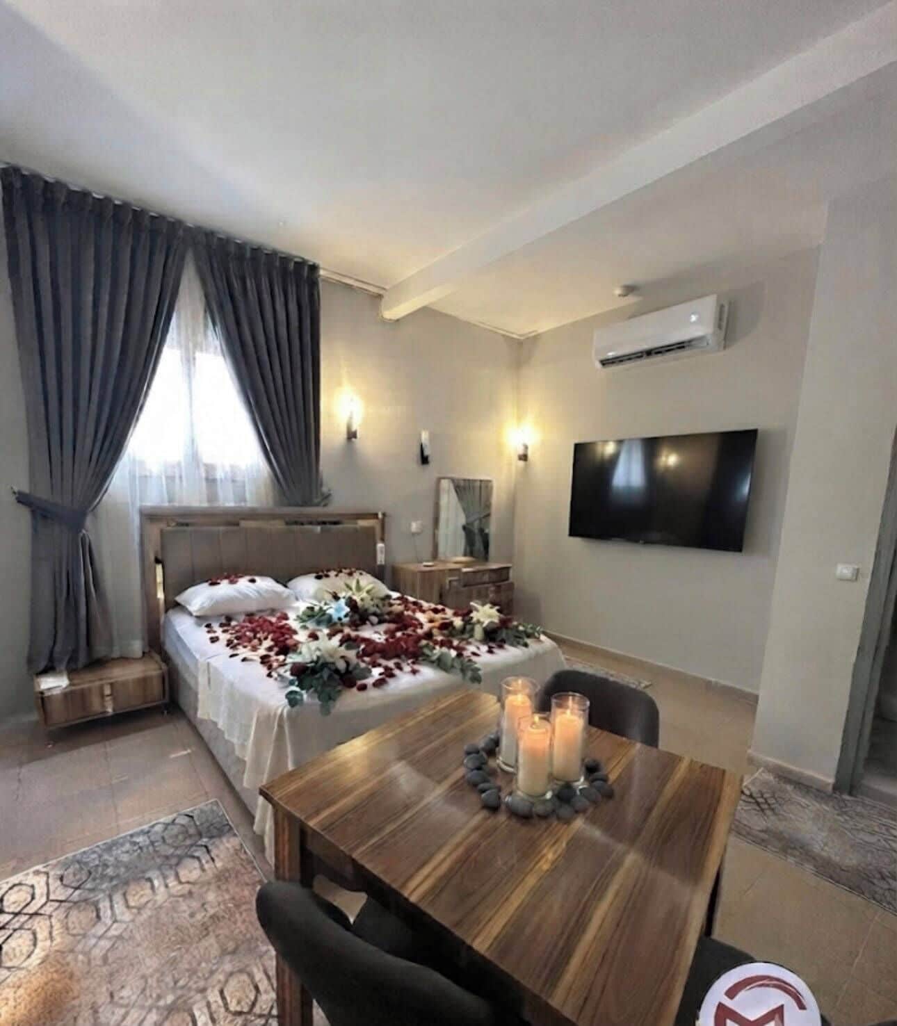 A hotel room with a bed, a dining table with candles, a TV mounted on the wall, and an air conditioning unit.