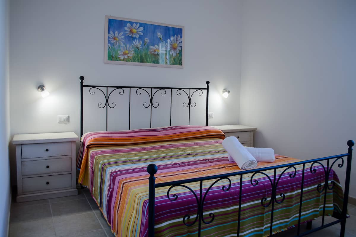 Un dormitori amb un llit, una tauleta de nit, un moble amb calaixos i un quadre a la paret.