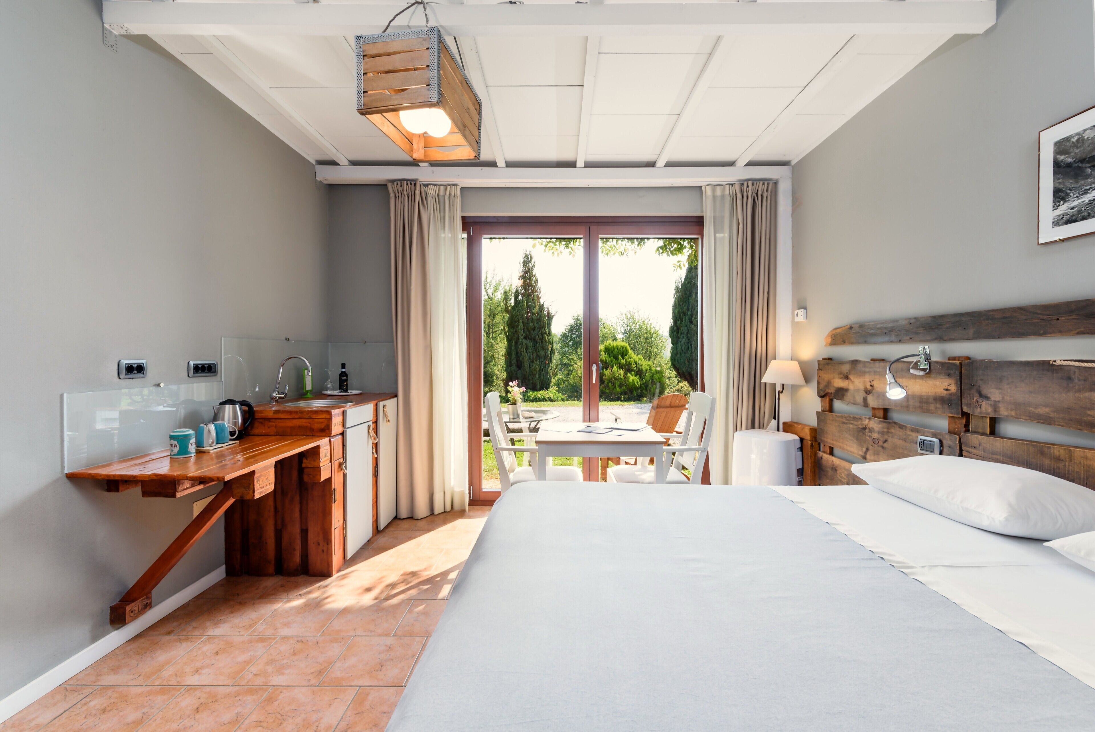 A bedroom with a large bed, a wooden desk, a window with curtains, and a view of greenery outside.