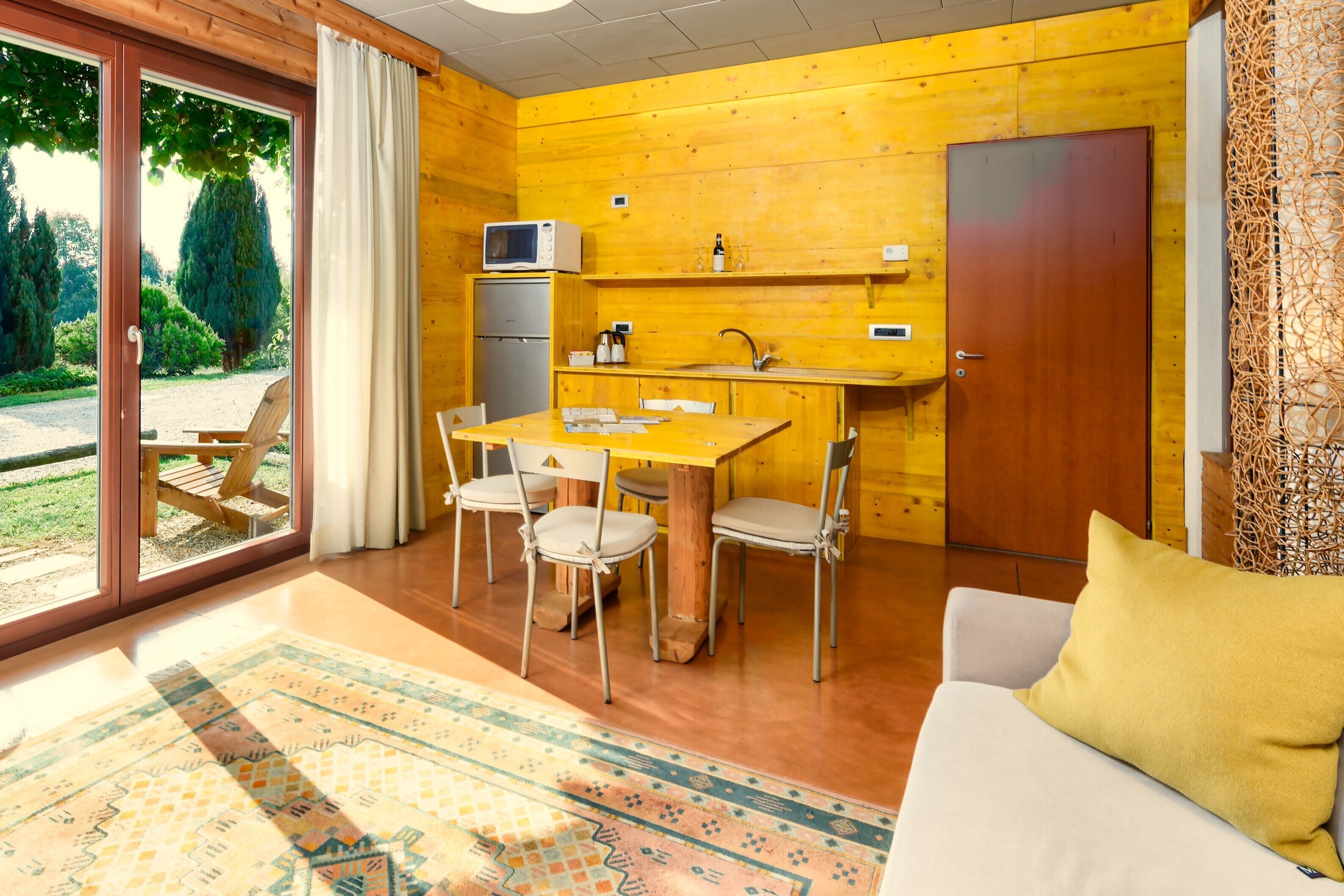 Een knus vertrek met houten details, een eettafel met stoelen, een bank en een groot raam met gordijnen.