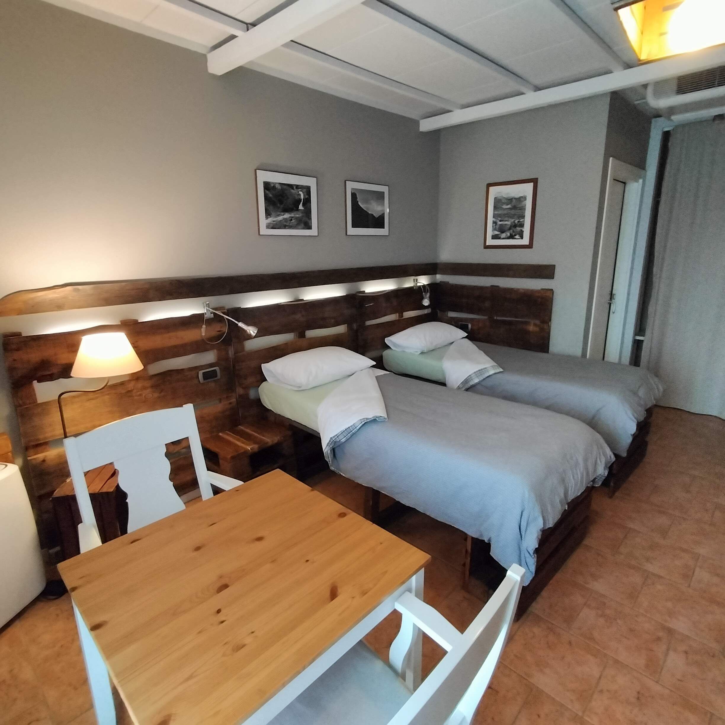 Een hotelkamer met een houten hoofdbord, twee enkele bedden, een houten tafeltje en een stoel. Er hangen afbeeldingen in lijsten aan de muur en op het nachtkastje staat een lamp.