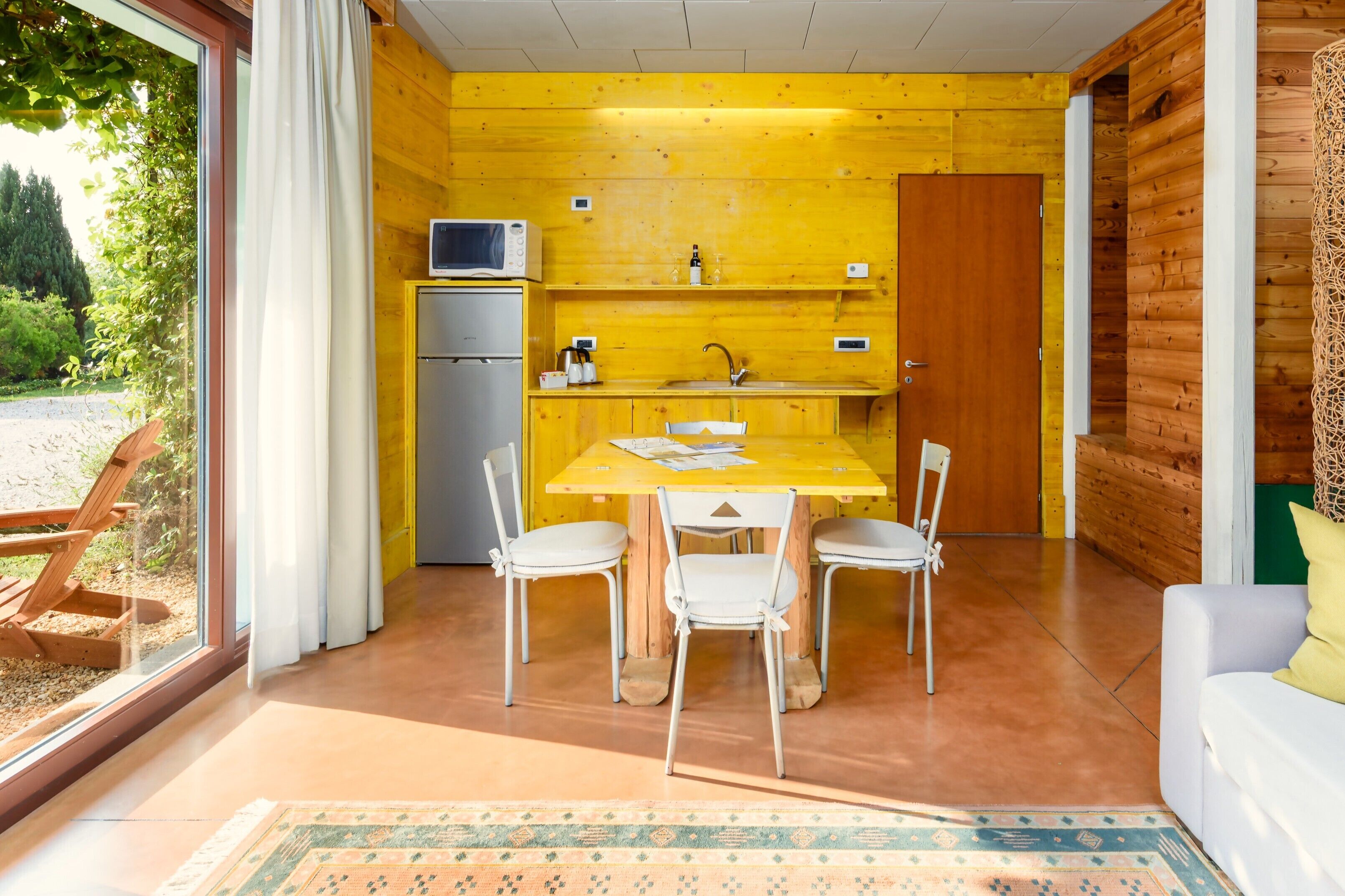 Cocina compacta con pared amarilla, un refrigerador, un microondas y un lavamanos. Hay una mesa de comedor con cuatro sillas y un tapete en el suelo.
