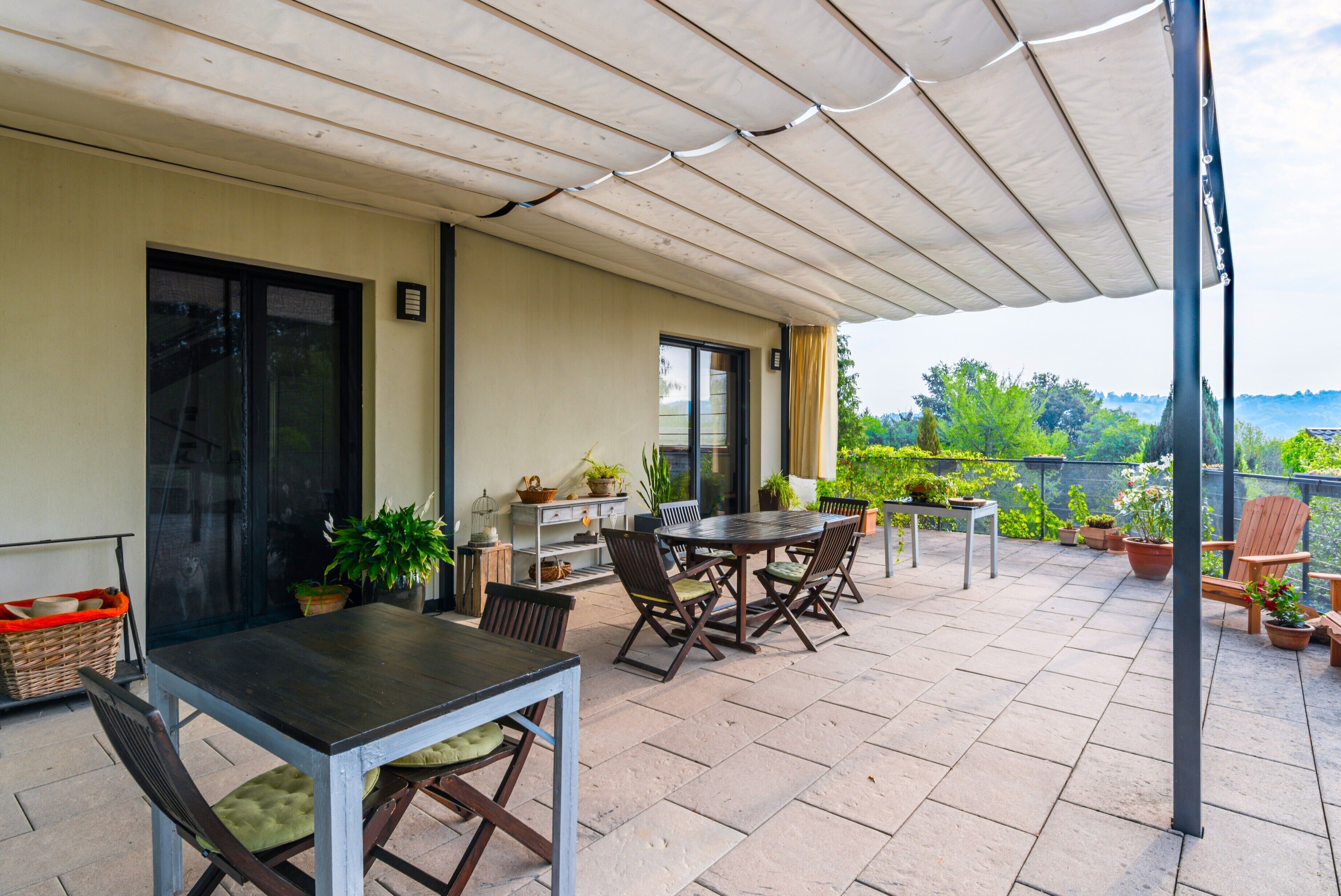 A covered outdoor patio with a table and chairs, potted plants, and a view of greenery.
