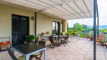 A covered outdoor patio with a table and chairs, potted plants, and a view of greenery.