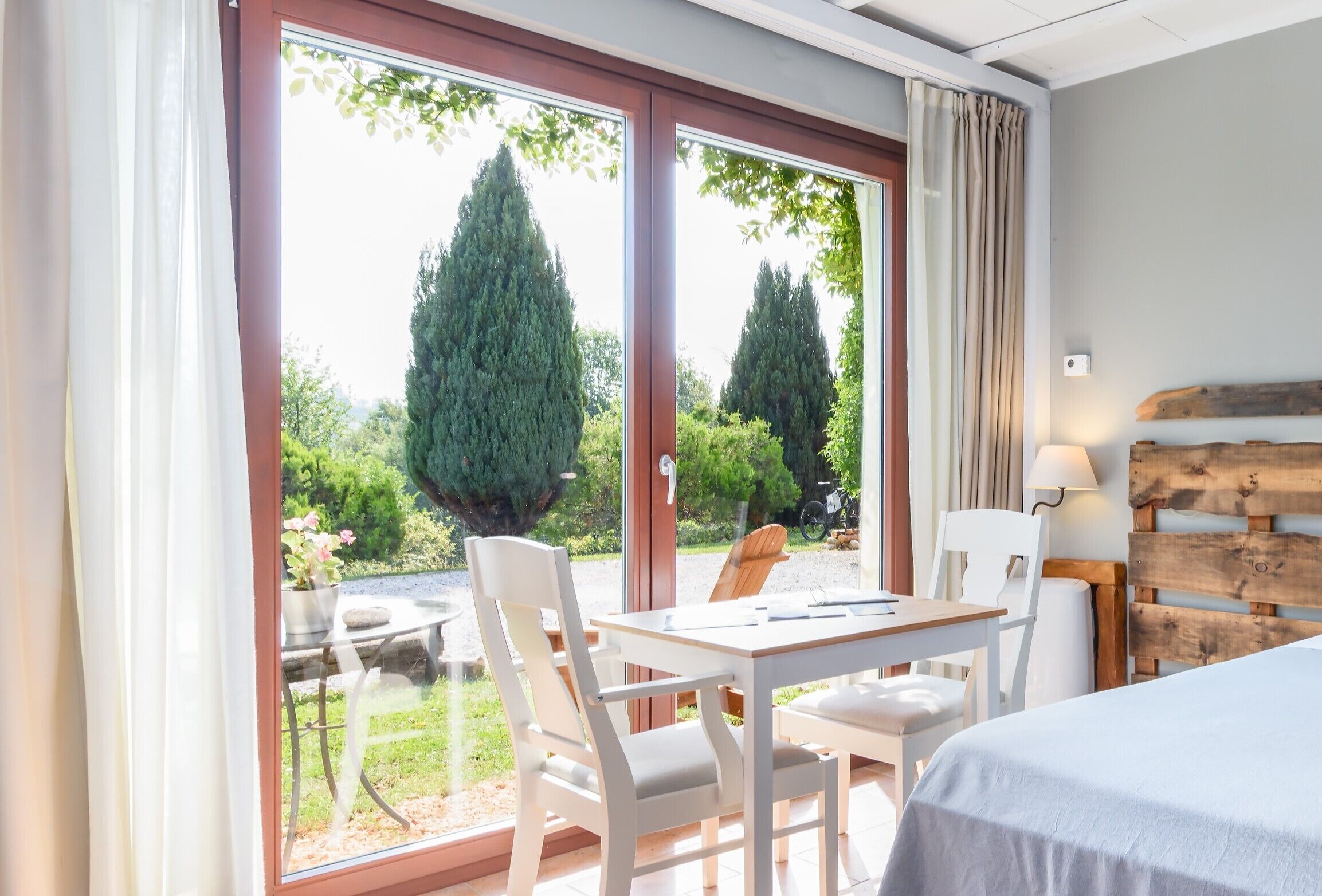 Una habitación con cama, una mesa con sillas, un ventanal con cortinas y vistas al exterior.