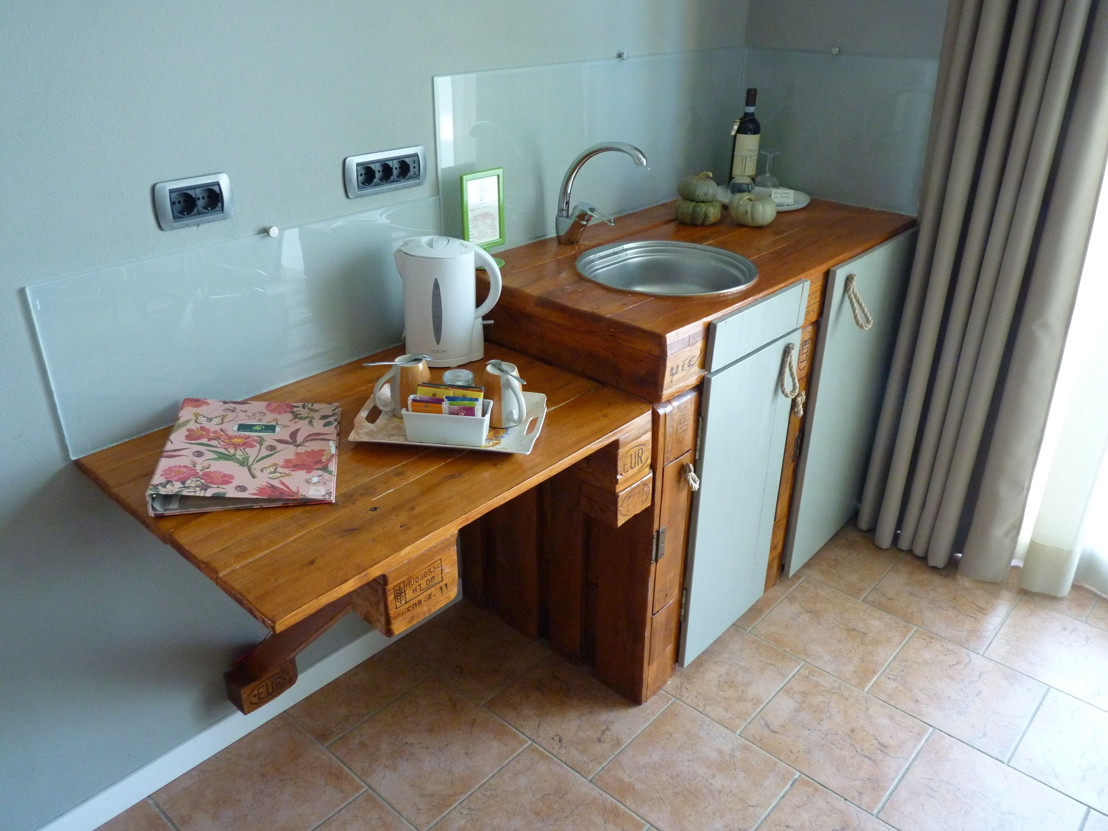 A kitchen with a wooden countertop, a sink, a kettle, a wine bottle, and a small window with curtains.