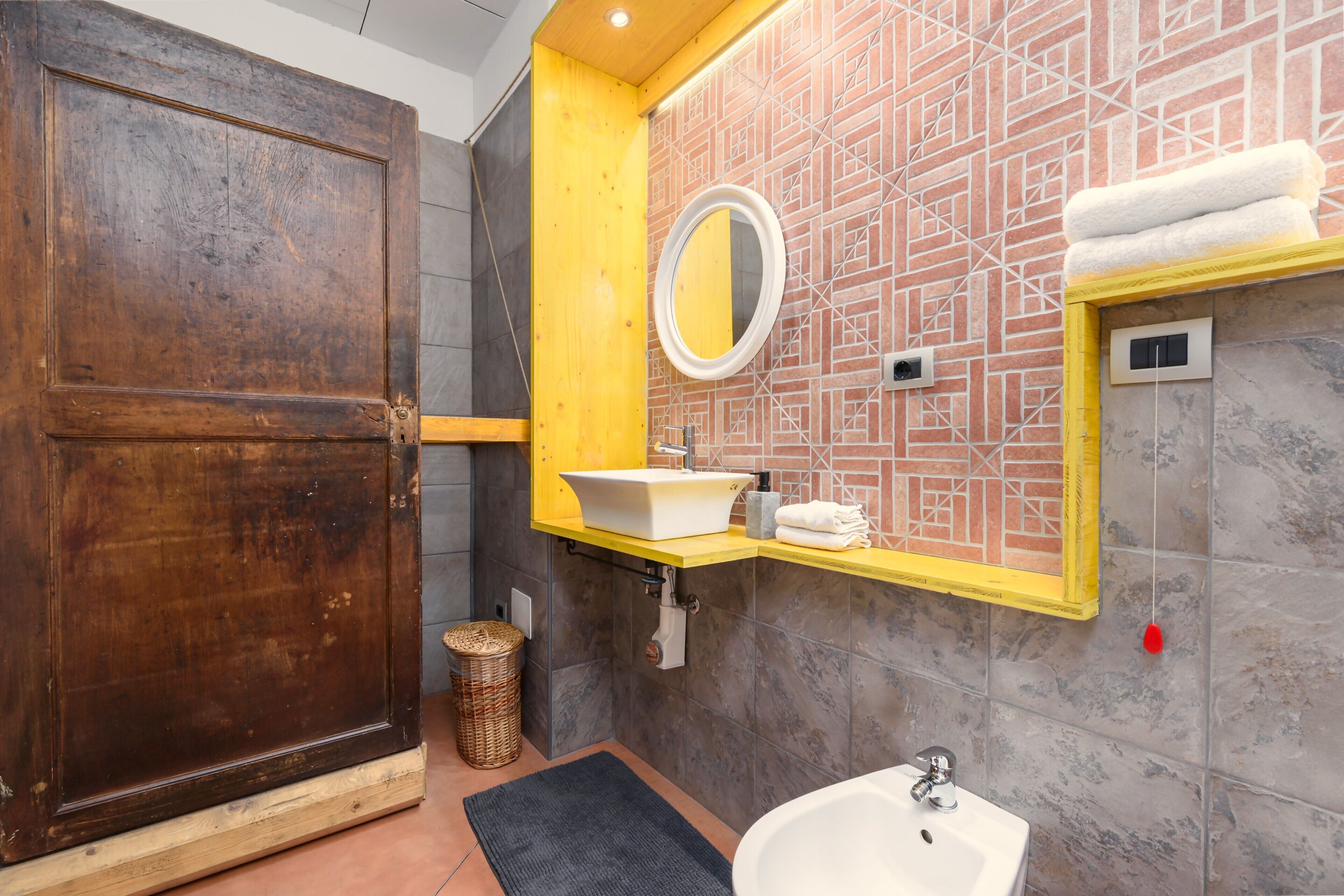 Un baño con puerta de madera, espejo con marco amarillo, lavabo blanco y un interruptor de luz rojo.