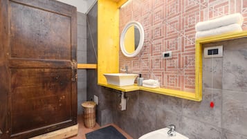 Un baño con puerta de madera, espejo con marco amarillo, lavabo blanco y un interruptor de luz rojo.