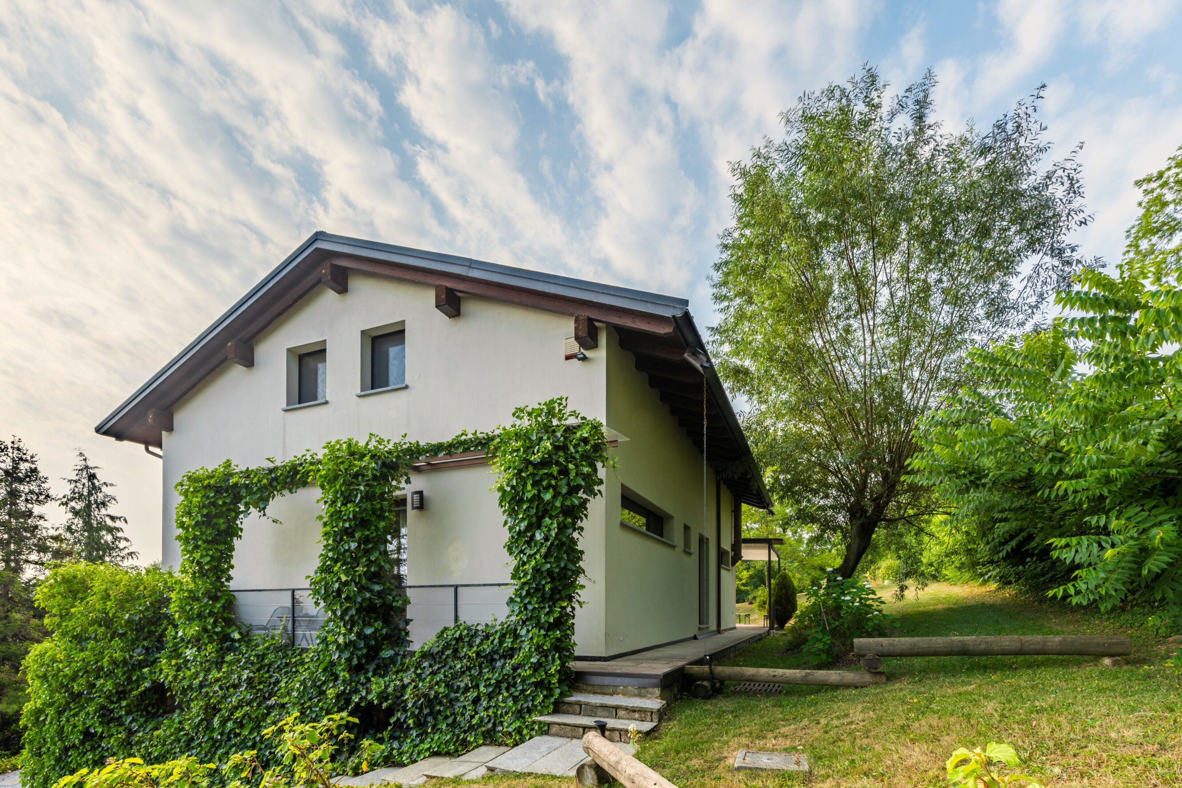 Ein zweigeschossiges Haus mit geneigtem Dach, einer Veranda und einem Garten mit verschiedenen Pflanzen und Bäumen.