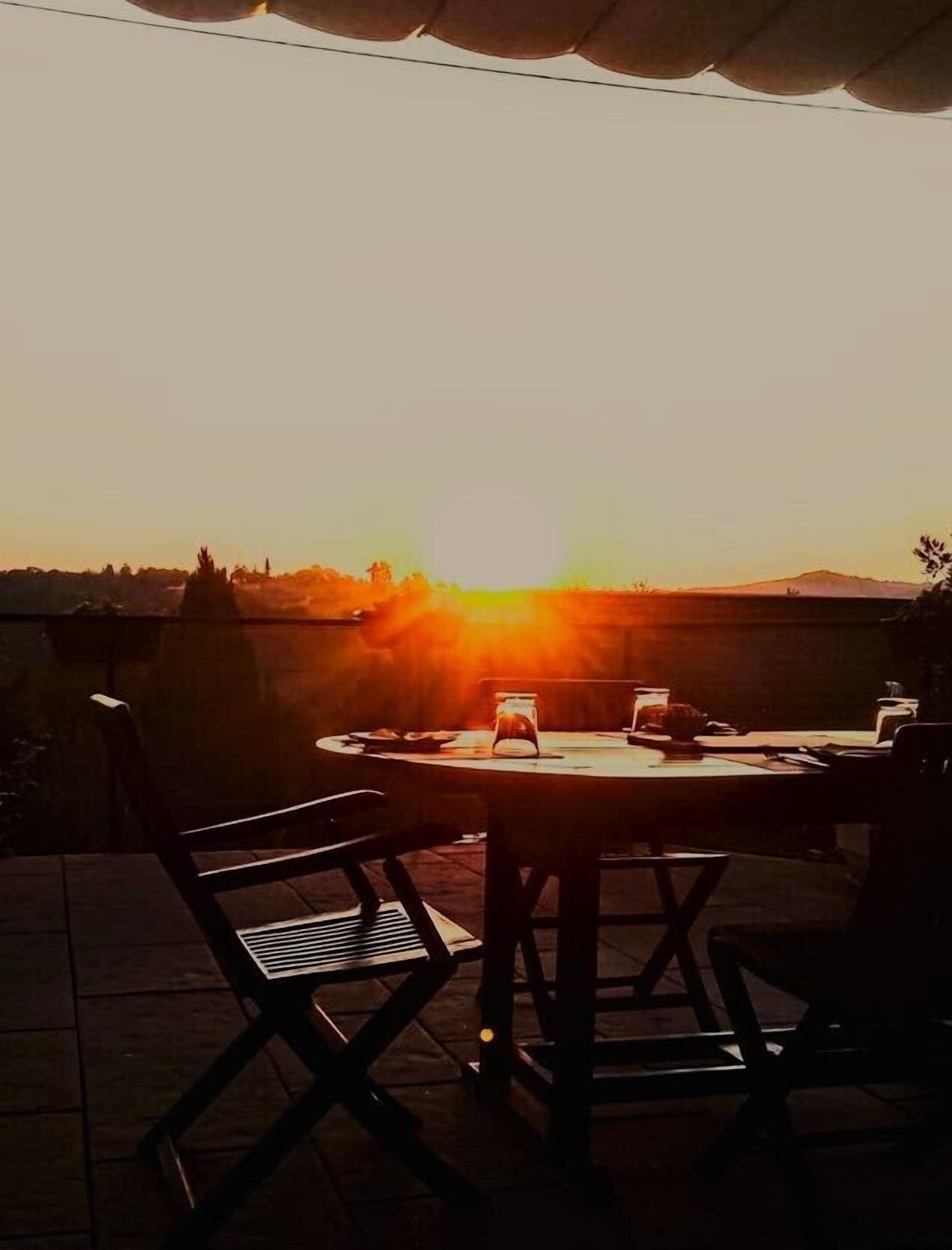Un patio al aire libre con una mesa redonda y sillas, con vista a una puesta de sol y una montaña distante.