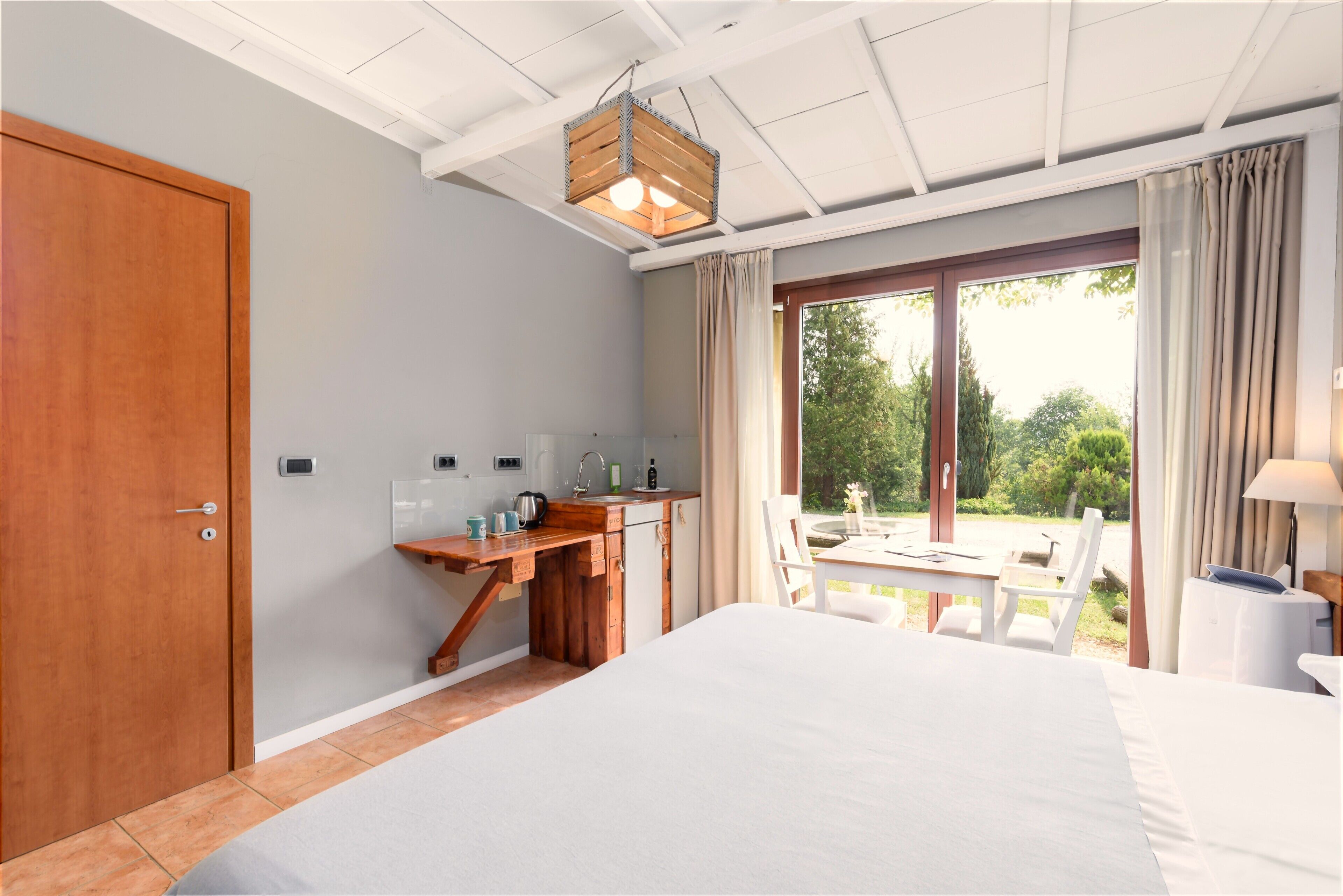 Quarto com cama, escrivaninha de madeira, janela com cortinas e vista para o verde lá fora.