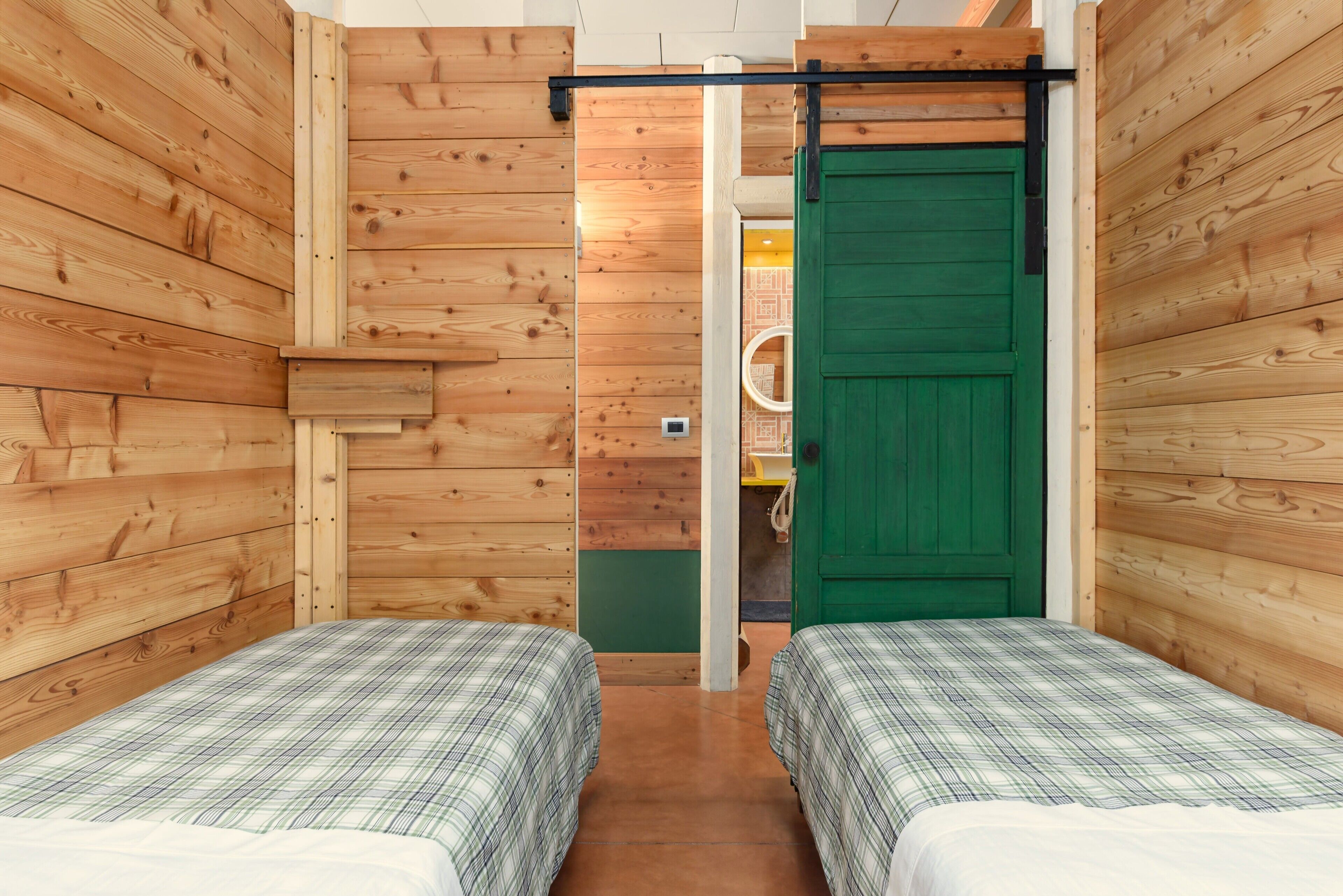 Una habitación con dos camas, paredes de madera, una puerta verde y un estante pequeño.