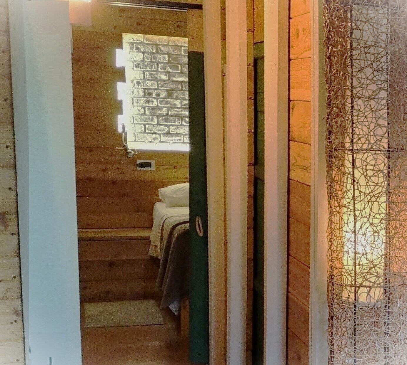 Ein schmaler Flur, der zu einem Raum mit Holzwand, kleinem Fenster und einem Bett mit weißem Kopfkissen und Decke führt.
