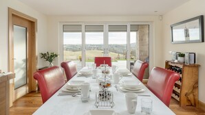 A dining area with a long table set for a meal, red chairs, a wine rack, and a view of the countryside through large windows. - The Queens View Luxury B&B (Linlithgow)