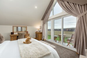 A bedroom with a large bed, a desk, a chair, and a view of a grassy field through large windows. - The Queens View Luxury B&B (Linlithgow)
