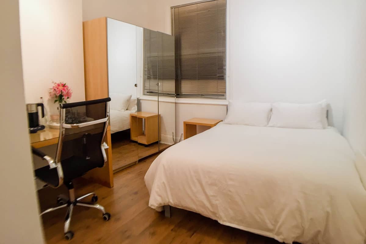A bedroom with a bed, a desk with a chair, a small table, and a window with blinds.